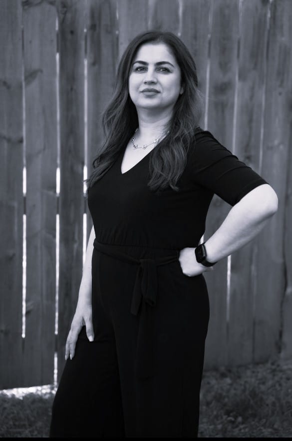 A black-and-white photo of a woman in a black dress standing confidently with one hand on her hip, against a wooden fence background.