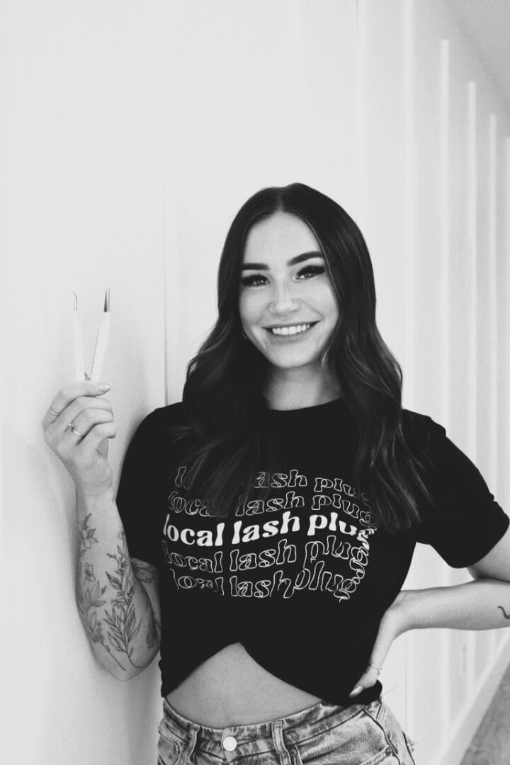 A woman smiles while holding a pair of tweezers in one hand. She is wearing a black shirt with text and jeans.