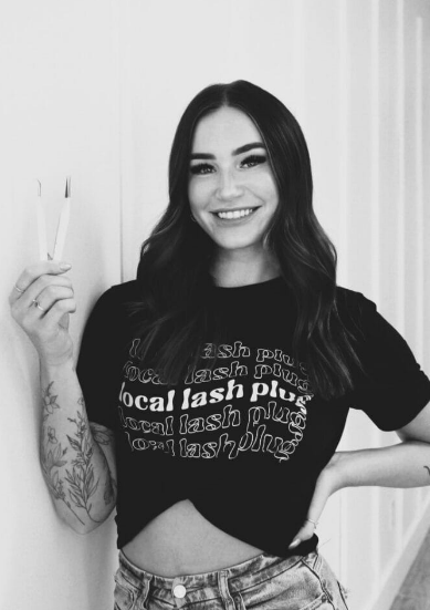 A woman smiles while holding a pair of tweezers in one hand. She is wearing a black shirt with text and jeans.